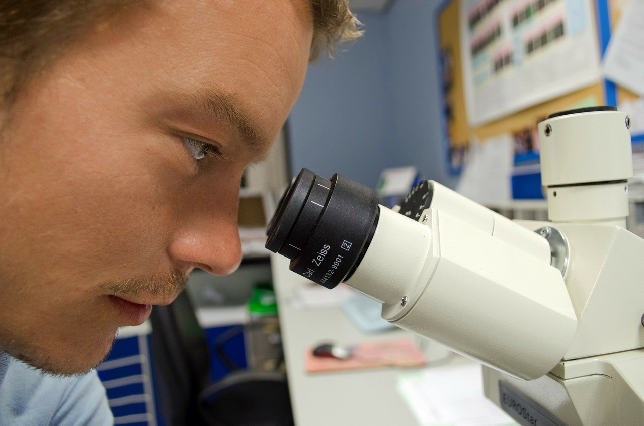 The image shows a man looking through a microscope. The man appears to be using the Carl Zeiss microscope.
