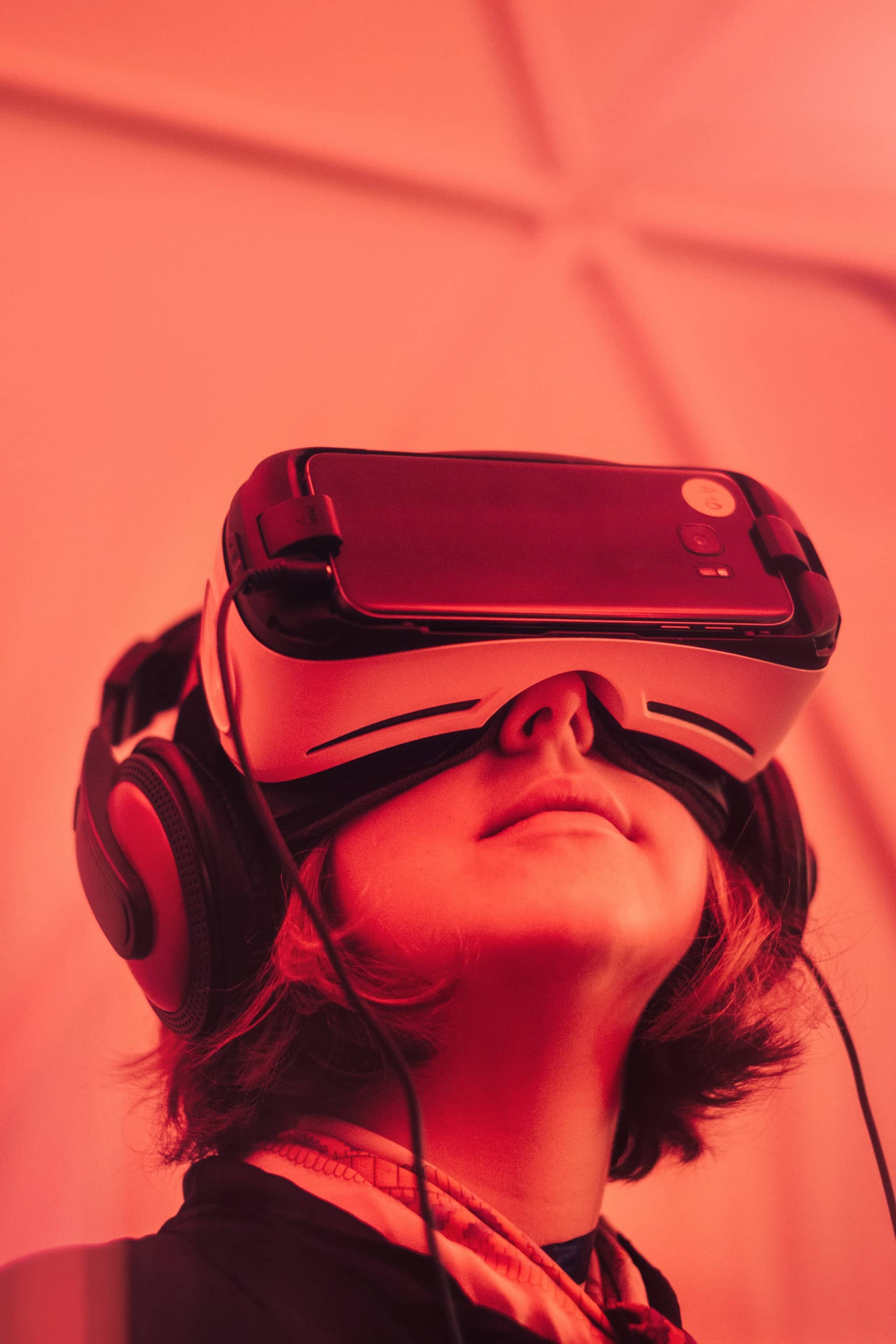 The photo is of a person wearing a virtual reality headset. The individual has a human face and is wearing red clothing and headphones. The virtual reality headset is considered a fashion accessory in this image, and the person appears to be listening to music.